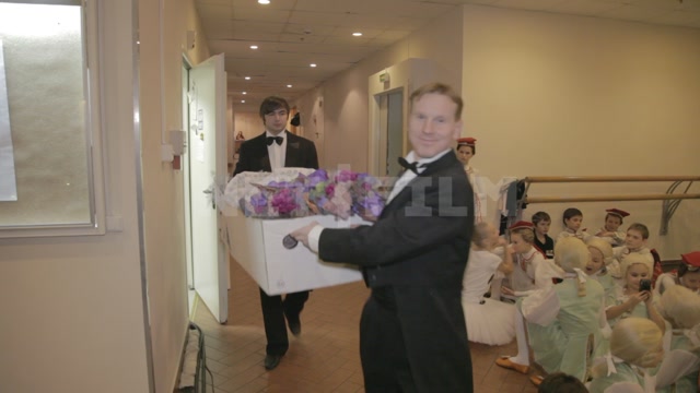 Men in tailcoats carry baskets of flowers and go backstage. The frame shows brass band musicians